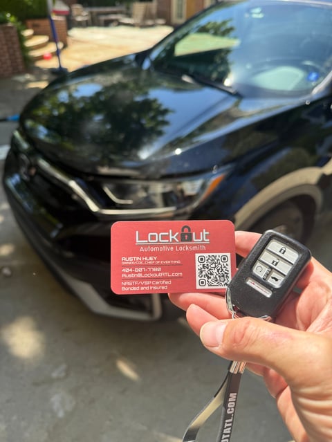 Hand holding a red Lockout automotive locksmith business card and car key fob in front of a dark sedan