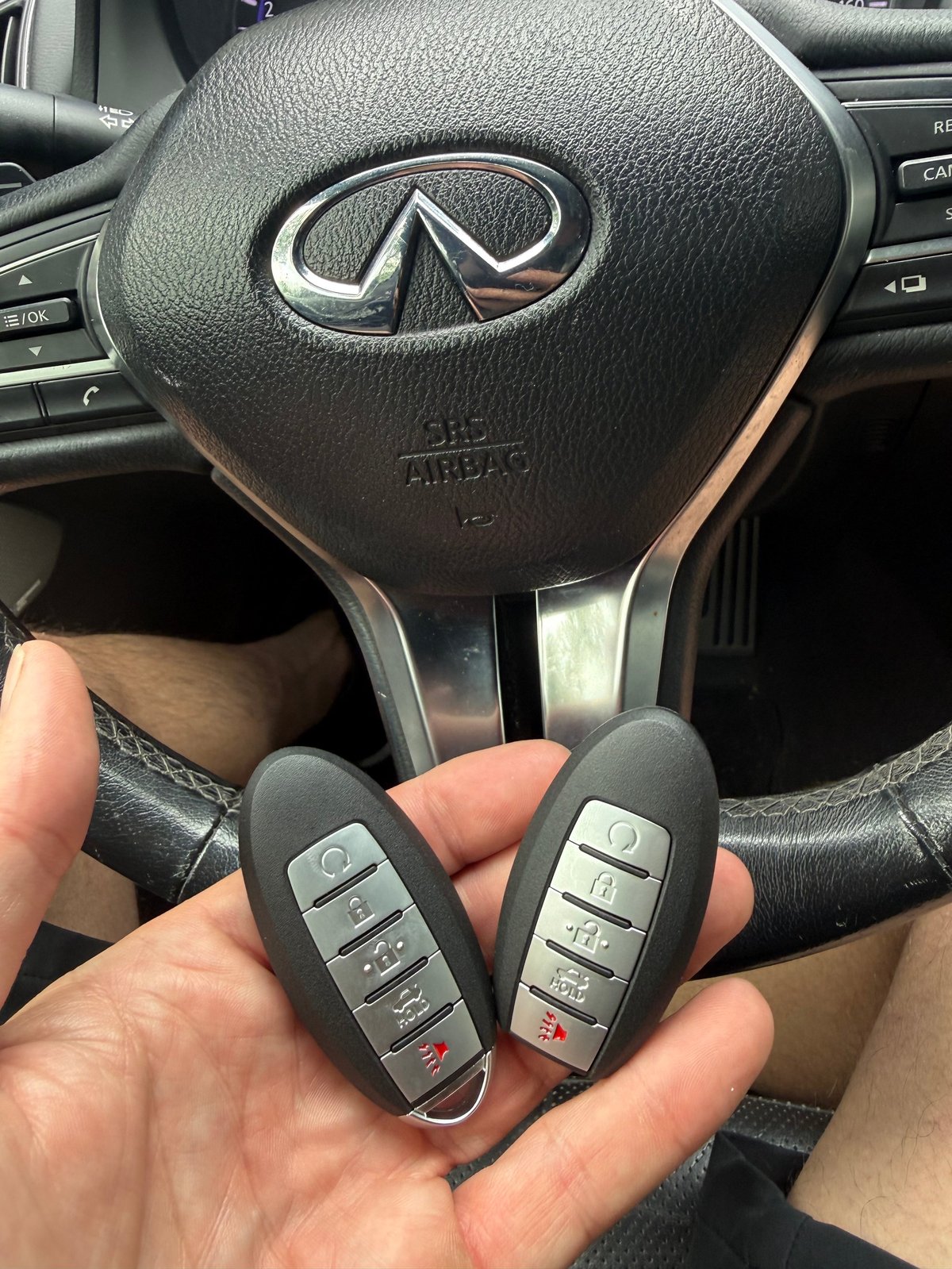 Person holding two Infiniti car key fobs in front of a steering wheel with Infiniti logo in a vehicle interior