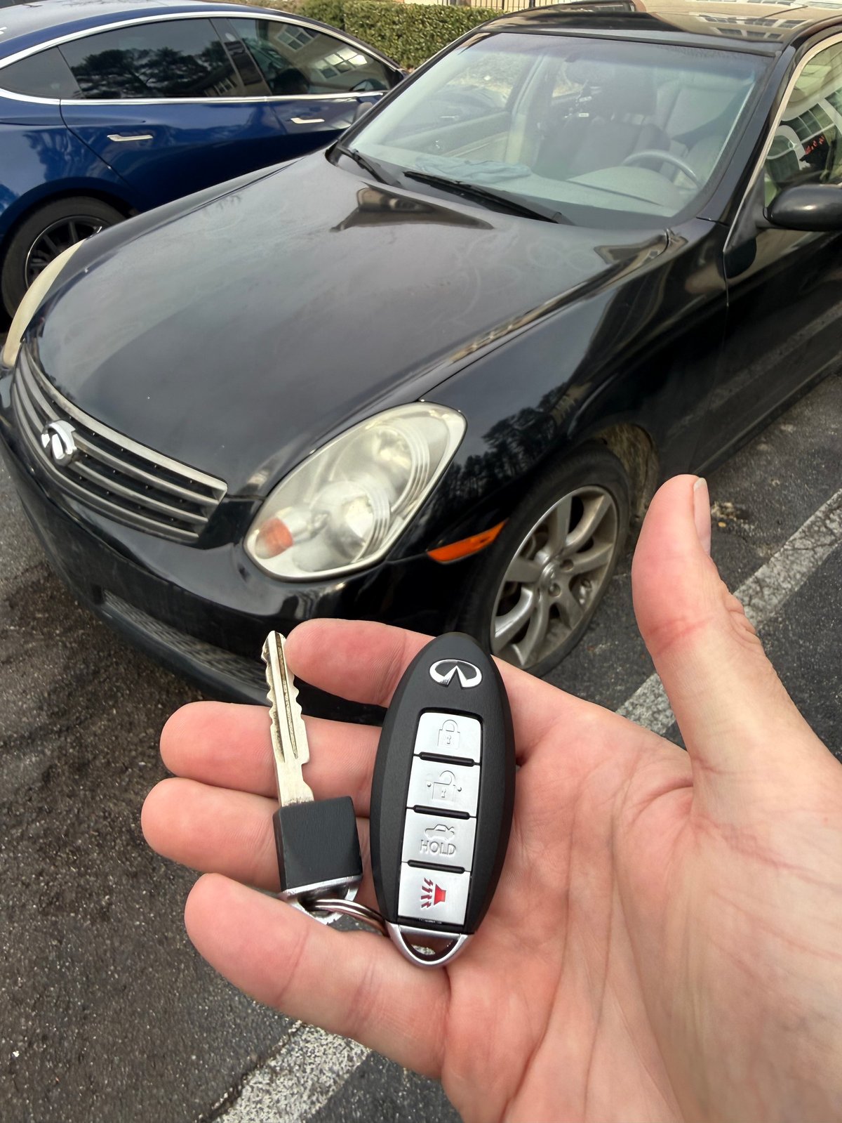 Hand holding a black Infiniti car key fob with a parking lot of vehicles in the background